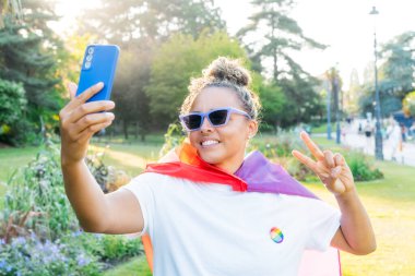 Gökkuşağı bayrağına sarılmış, LGBT topluluğunun sembolü olan genç Latin mutlu kadın, bir şehir parkındaki bir gurur kutlamasında cep telefonuyla selfie çekiyor. İnsan hakları, eşitlik, LGBT, Özgürlük ve Mutluluk