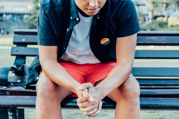 Cropped no face sad frustrated LGBTQ person with rainbow badge on t-shirt sitting on park bench alone. Concept of loneliness, sadness and pain for homosexual discrimination. Mental crisis, depression.