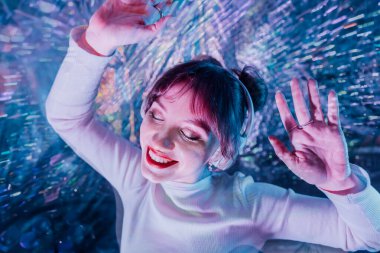 Mutlu, gülümseyen, gözleri kapalı, beyaz elbiseli ve kulaklıklı dans eden bir kadın. Müzik aşığı. Beyaz elbiseli parti, sessiz disko. Parlak arka planda iyi eğlenceler. Seçici odak.