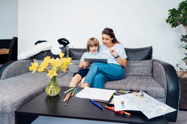 Anne ve oğlu dijital tablet kullanarak kanepede yatıyor. Mutlu anne ve küçük çocuk video izlemek, kitap okumak, video oyunu oynamak, online uygulama kullanmak, evde birlikte çalışmak için dokunmatik ekran tableti kullanıyorlar..