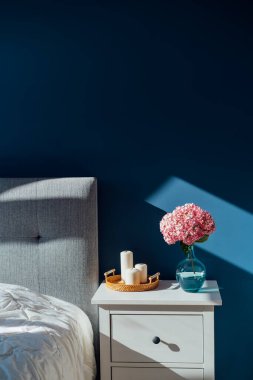 Koyu renkli modern yatak odası. İçi lacivert duvarlarla, ev dekoruyla dolu. Gri yatak başlıklı yatak, beyaz battaniye, komodin, pembe ortanca çiçekli vazo, mumlar. Dikey