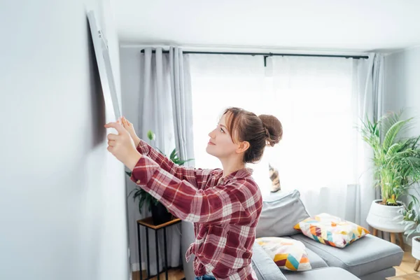 stock image Side view young woman putting painting, mockup picture frame on the wall. Casual lady taking care of coziness in her new stylish apartment. Young female decorating room. Moving and renovate house.