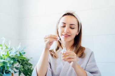 Young woman in bathrobe looking in the mirror, holding dropper with hyaluronic acid serum close to face and smiling. Skin hydrating. Cosmetic spa procedures. Beauty self-care at home. Selective focus.