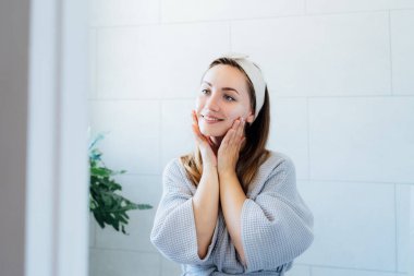 Genç, gülümseyen bir kadın krem maske takıyor ve banyoda kendine elle masaj yapıyor. Cilt bakımı için doğal prosedürler. Evde güzellik tedavisi, gençler için yüz jimnastiği. Seçici odak