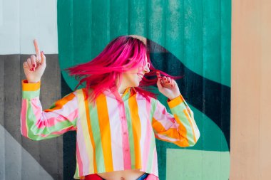 Parlak pembe saçlı, güneş gözlüklü, graffiti arka planında çizgili tişörtlü duygusal genç bir kadın elleriyle işaret ediyor ve başını yana çeviriyor. Uçan saç. Seçici odak