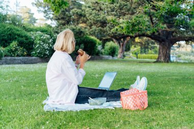 Side view middle-aged woman sitting on green grass lawn, eating healthy lunch and working on laptop. Working remotly, having picnic during break. Balanced diet lunch box with salad and sanwich