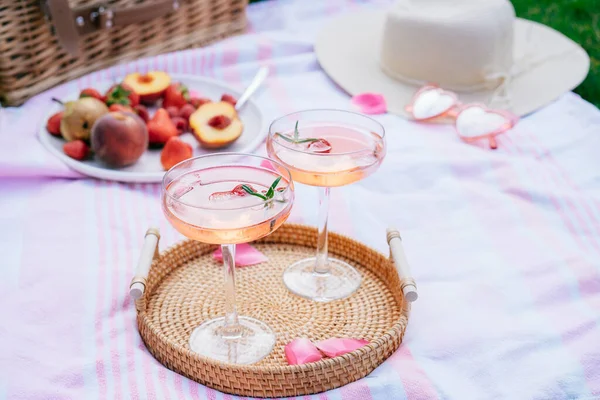 Pink drinks, cocktail with ice, raspberry, rosemary. Two glasses with martini, champagne, cider, lemonade on the blanket with fruit plate, picnic basket. Cozy summer picnic on nature. Selective focus.