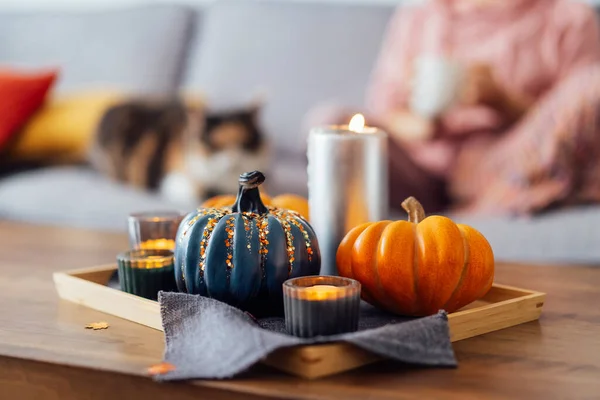 Autumn, fall cozy mood composition for hygge home decor. Small pumpkins, burning candles on tray with gray napkin on the coffee table with resting woman with cat in the living room. Selective focus.