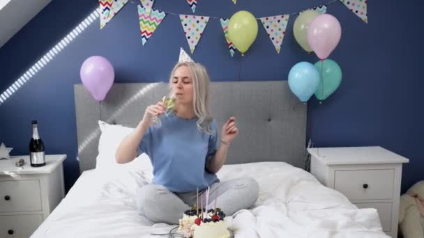 穿着睡衣的快乐女人 戴着派对帽在床上跳舞 手里拿着香槟酒杯 床上拿着喜庆的蛋糕 是时候在家里庆祝了生日快乐的概念 — 图库视频影像