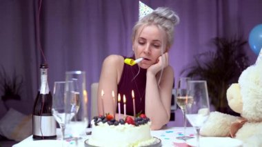 Sad attractive woman in party cap blowing in birthday pipe while celebrating birthday at home, sitting alone at served table with cake, keeping hand under chin, looking away, dreaming. Bored party.