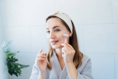 Bornozlu genç bir kadın aynaya bakıyor ve banyoda yeşim taşıyla yüz masajı yapıyor. Cilt bakımı için doğal prosedürler kavramı. Ev Güzeli kendi kendine bakım tedavisi. Seçici odak.