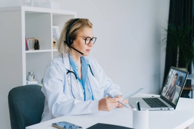 Beyaz önlüklü orta yaşlı doktor ve video konferans kulaklığı dizüstü bilgisayarında hastaya online video görüşmesi yapıyor. Ev sağlığı tedavisi için teletıp konsepti. Uzaktan kumandalı tıbbi randevu