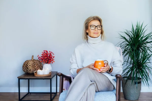 Orta yaşlı bir kadın kabak şeklinde bir bardak sıcak içecekle gevşiyor. Skandal stili Hygge tarzı dekorlu bir iç mekan. Rüya gören bayan, stres olmadan sakin havanın tadını çıkar, yalnız olmanın tadını çıkar. Sıcak sonbahar.