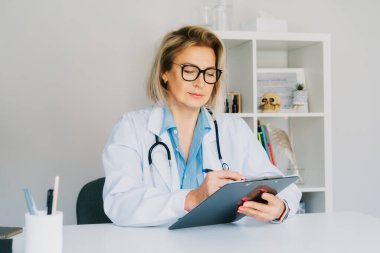 Kendinden emin orta yaşlı kadın doktor hemşire, beyaz önlüklü kadın doktor steteskoplu pratisyen hekim hastane ofisinde çalışıyor ve randevu sırasında not alıyor.