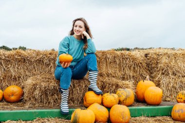 Gülen genç bir kadın saman balyalarının üzerinde oturuyor ve elinde balkabakları tutuyordu. Tarım çiftliğindeki Şükran Günü ve Cadılar Bayramı dekorasyonu için en iyisini seçiyorum. Balkabağı hasadı. Sonbahar şenlik havası..