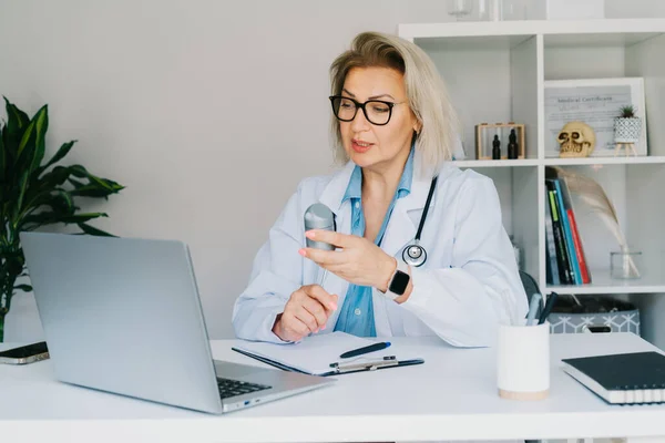 Confident mature middle aged woman doctor shows jar of pills to laptop in clinic office interior. Video call online with patient, treatment remote, pharmacy, advice, medical healthcare. Telemedicine.