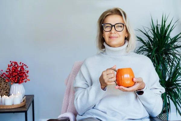 Orta yaşlı bir kadın kabak şeklinde bir bardak sıcak içecekle gevşiyor. Skandal stili Hygge tarzı dekorlu bir iç mekan. Rüya gören bayan, stres olmadan sakin havanın tadını çıkar, yalnız olmanın tadını çıkar. Sıcak sonbahar