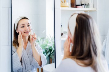 Bornozlu genç bir kadın aynaya bakıyor ve mikro iğneyle yüz masajı yapıyor. Cilt bakımı için doğal prosedürler kavramı. Ev Güzeli kendi kendine bakım tedavisi. Seçici odak