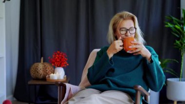 Orta yaşlı bir kadın kabak şeklinde bir bardak sıcak içecekle gevşiyor. Skandal stili Hygge tarzı dekorlu bir iç mekan. Rüya gören bayan, stres olmadan sakin havanın tadını çıkar, yalnız olmanın tadını çıkar. Sıcak sonbahar.
