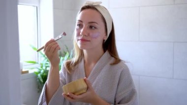 Young woman in bathrobe looking in the mirror and applying natural cosmetic clay mask on her face in bathroom. Cosmetic procedures for teens skin care at home. Beauty self-care. Selective focus.