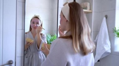 Young woman in bathrobe looking in the mirror and applying natural cosmetic clay mask on her face in bathroom. Cosmetic procedures for teens skin care at home. Beauty self-care. Selective focus.