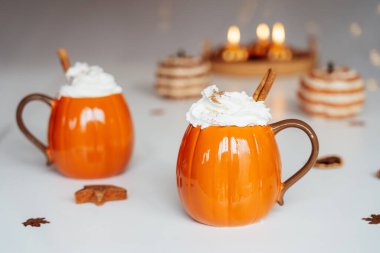Balkabağı şekilli krem şantili ve tarçınlı bardaklar beyaz masada sonbahar dekoru ile. Baharatlı balkabaklı latte. Şükran Günü için sıcak sonbahar içeceği ya da evde Cadılar Bayramı tatili.
