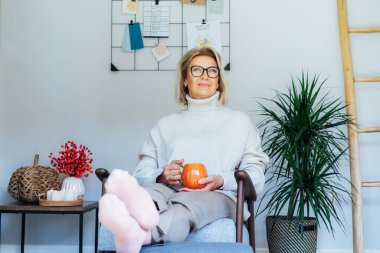Orta yaşlı bir kadın kabak şeklinde bir bardak sıcak içecekle gevşiyor. Skandal stili Hygge tarzı dekorlu bir iç mekan. Rüya gören bayan, stres olmadan sakin havanın tadını çıkar, yalnız olmanın tadını çıkar. Sıcak sonbahar