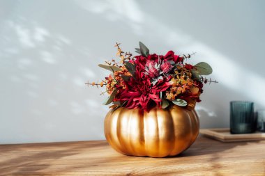 Altın kabak vazosundaki parlak yapay çiçeklerden oluşan sonbahar buketi, arka planda gölgeler olan beyaz duvarlı ahşap konsoldaki mumlar. Hygge rahat bir ev atmosferi için sonbahar dekoru