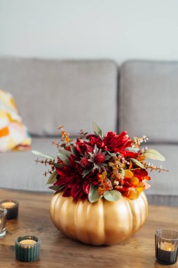 Altın kabak vazosundaki parlak yapay çiçeklerden oluşan sonbahar buketi oturma odasının içindeki ahşap sehpada mumlar yakıyor. Hygge 'nin evi rahat bir ev atmosferi için sonbahar dekorasyonu. Dikey.