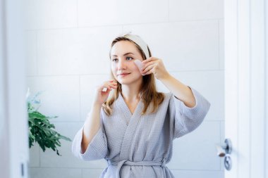 Bornozlu genç bir kadın aynaya bakıyor ve tuvalette gua sha roller ile yüz masajı yapıyor. Cilt bakımı için doğal prosedürler kavramı. Ev Güzeli kendi kendine bakım tedavisi. Seçici odak