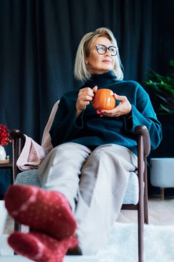 Orta yaşlı bir kadın kabak şeklinde bir bardak sıcak içecekle gevşiyor. Skandal stili Hygge tarzı dekorlu bir iç mekan. Rüya gören bayan, stres olmadan sakin havanın tadını çıkar, yalnız olmanın tadını çıkar. Sıcak sonbahar