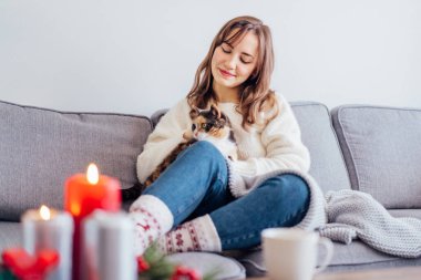 Hygge ev dekorasyonu için Noel kompozisyonu. Yanan mumlar, sehpadaki tepside çam ağacı dalları sıcak çay içen ve kanepede kedi besleyen dinlenen bir kadınla. Seçici odak