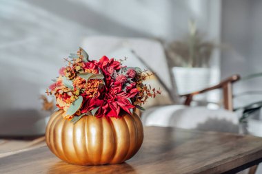 Ahşap sehpanın üzerinde, modern oturma odası arka planına sahip altın bir kabak vazosundaki parlak yapay çiçeklerden oluşan sonbahar buketi. Hygge rahat bir ev atmosferi için sonbahar dekoru