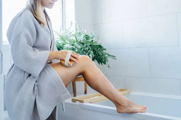 Young woman in bathrobe doing scrubbing massage with dry brush before morning shower in light colored bathroom. Anti-cellulite treatment, lymphatic drainage massage. Women beauty. Selective focus.