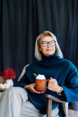 Orta yaşlı kadın bir bardak sıcak içecekle gevşiyor. İç dekorasyonları sonbahar modunda. Rüya gören bayan, stres olmadan sakin havanın tadını çıkar, yalnız olmanın mutluluğu. Evde rahat bir sonbahar.