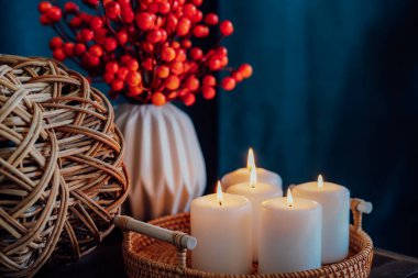Hygge ev dekoru için sonbahar havası kompozisyonu. Hasır balkabağının bir parçası, tepside mum yakmak ve tahta sehpanın üzerinde, koyu arkaplanlı vazoda mevsimlik çiçekler. Seçici odak.