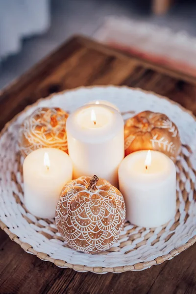 Close up autumn fall cozy mood composition for hygge home decor. Orange pumpkins decorated with mandalas, white burning candles on wicker plate on the coffee table. Selective focus. Vertical card