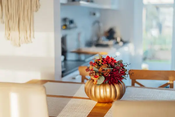 Autumn bouquet of bright artificial flowers in a golden pumpkin vase on a kitchen table counter with white modern kitchen background. Autumn home decor for Thanksgiving and Halloween holiday