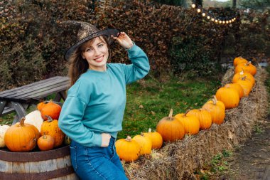 Cadı şapkalı gülümseyen genç kadının portresi balkabağı arka planında poz veriyor. Kadın, tarım çiftliğinde Şükran Günü ve Cadılar Bayramı dekorasyonu için en iyi balkabaklarını seçiyor. Sonbahar şenlik havası.