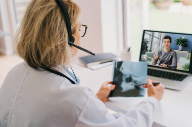Beyaz önlüklü orta yaşlı doktor ve video konferans kulaklığı dizüstü bilgisayarında hastaya online video görüşmesi yapıyor. Ev sağlığı tedavisi için teletıp konsepti. Uzaktan kumandalı tıbbi randevu