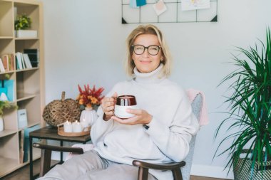 Gülümseyen orta yaşlı bir kadın sıcak kahve içmekten zevk alıyor ve dağınık stil hygge iç mekanında sonbahar modunda rahatlıyor. Rüya gören bir kadın, stressiz sakin bir ruh hali, yalnız olmak. Sıcak sonbahar.