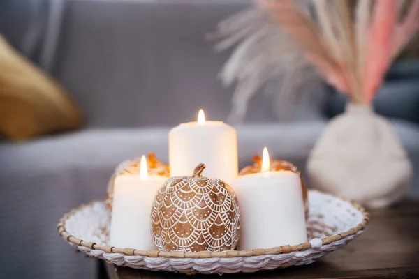 Autumn fall cozy mood composition for hygge home decor. Orange pumpkins decorated with mandalas, white burning candles on wicker plate on the coffee table in the living room. Selective focus.