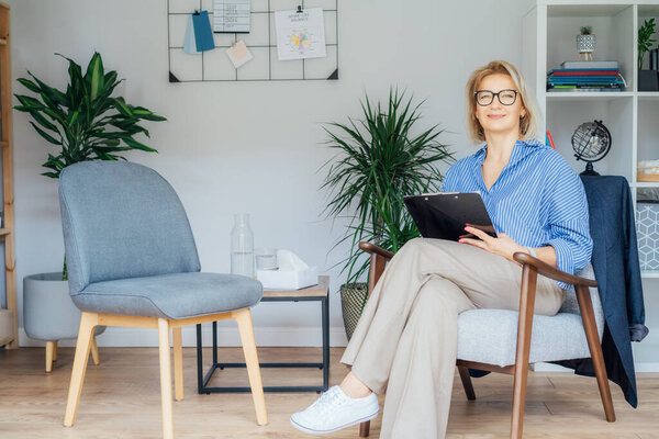 Professional Psychotherapy. Successful female Psychologist Smiling To Camera Sitting On armchair In Office. Mature 50s middle-age professional portrait of teacher, coach, mentor, therapist, counselor.