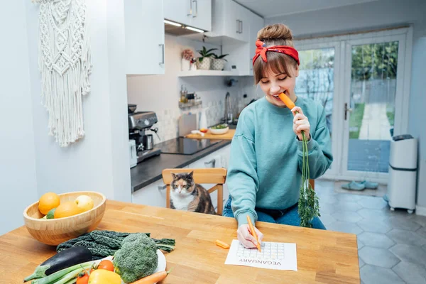 Young Woman Filling Veganuary Calendar While Eating Fresh Carrot Sitting — Stock Photo, Image