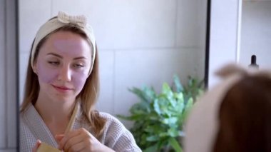 Young woman in bathrobe looking in the mirror and applying natural cosmetic clay mask on her face in bathroom. Cosmetic procedures for teens skin care at home. Beauty self-care. Selective focus.