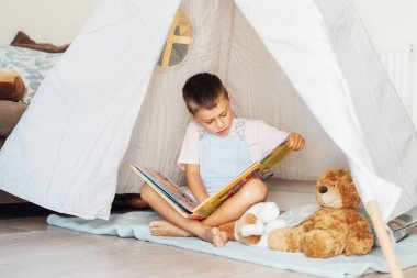 Sevimli küçük anaokulu çocuğu oyuncaklarına kitap okuyor rahat çocuklar çadırında oturuyor evdeki oturma odasında. Çocuk kendini eğlendiriyor. Ev çocukları etkinliği.