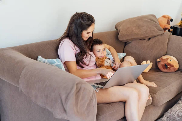 Young Mother and son on the sofa using laptop. Happy mom and little boy hugging and watching videos, reading books, playing video game, using online app, studying together at home. Family time.