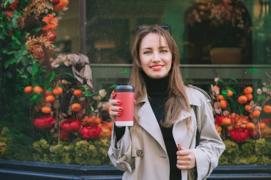 Palto giymiş genç bir kadının portresi yeniden kullanılabilir kahve fincanının içinde içki içiyor ve sonbahar tatili için vitrinleri dekore edilmiş şekilde caddede yürüyor. Mutlu sonbahar konforu, mevsimlik moda eğilimi