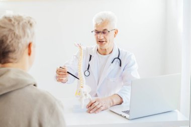 Orta yaşlı erkek doktor bir kalemle insan omurga modelini işaret ediyor ve hastanın klinikteki sorununu anlatıyor. Omurga sağlığı ve sırt ağrısı tedavisi konsepti. Seçici odak, boşluğu kopyala.