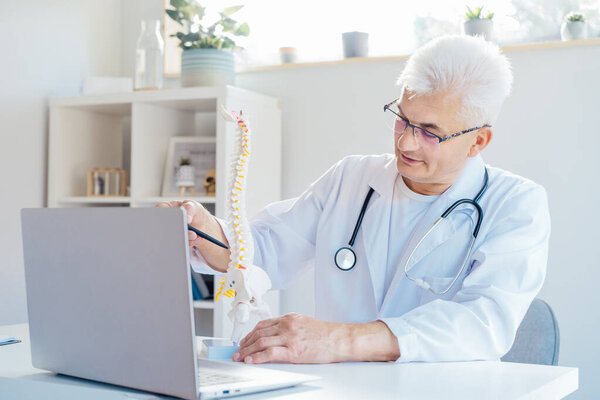 Doctor using laptop for telemedicine with patient at hospital. Osteopath makes online video call consult, pointing on human spine model. Spinal healthcare and back pain treatment. Remote appointment.